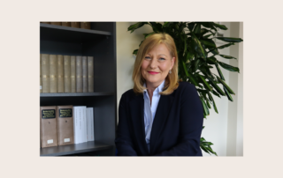 sharon wearing a shirt and blazer next to a book shelf, smiling.